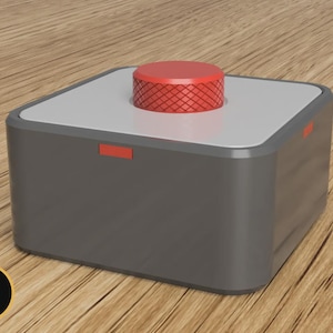May include: A grey square box with a red button on top. The button has a textured surface. A small red rectangle is on the side of the box. The box is on a wooden surface.