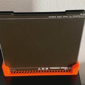 Könnte beinhalten: Eine schwarze, rechteckige beheizte Stahlplatte mit weißem Text, die von einer orangefarbenen Kunststoffbasis getragen wird. Der Text lautet "Heatbed Steel Sheet by PRUSA3D.COM" und "ORIGINAL PRUSA by Josef Prusa."