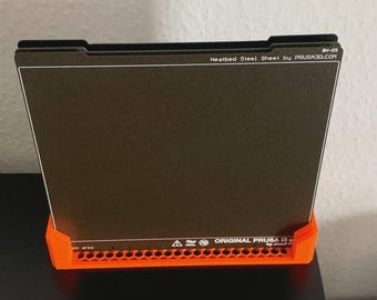 May include: A black rectangular heated steel sheet with white text, supported by an orange plastic base. The text reads "Heatbed Steel Sheet by PRUSA3D.COM" and "ORIGINAL PRUSA by Josef Prusa."