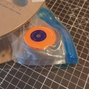 May include: An orange air valve with the text "Air Valve" in blue is sealed inside a clear plastic bag. The bag is partially visible, with a blue seal. The background includes a cutting mat and a portion of a brown cardboard roll.