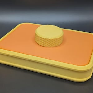 Könnte beinhalten: Ein rechteckiger Behälter in Gelb und Orange. Der Boden und der Rand sind gelb, während das Innere orange ist. Ein kleines, gelbes, zylindrisches Objekt mit einem Rautenmuster befindet sich oben. Wahrscheinlich zur Aufbewahrung.