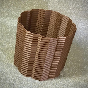 May include: A brown, textured, 3D-printed container with a unique, geometric design. The container has a wavy top edge and a series of angled, horizontal lines that create a layered effect. The container is made of a light brown material.