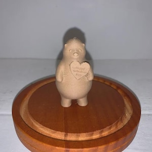 Puede incluir: Pequeña figura de oso beige sosteniendo un letrero en forma de corazón que dice "Happy Mother's Day". El oso está sobre una base redonda de madera con acabado marrón oscuro.