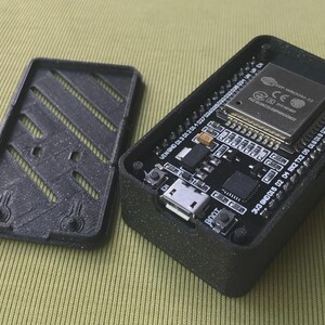May include: Black rectangular electronic device with an open lid, revealing a circuit board with various components. The lid has a slotted design. The device is on a light green surface. The circuit board has the text "BOOT" and "EN".