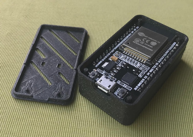 May include: Black rectangular electronic device with an open lid, revealing a circuit board with various components. The lid has a slotted design. The device is on a light green surface. The circuit board has the text "BOOT" and "EN".