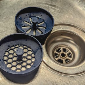 Op de afbeelding: Twee donkerblauwe gootsteenzeven met een honingraat- en stervormig patroon, naast een roestvrijstalen gootsteenafvoer. De zeven zijn van plastic.