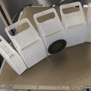 May include: A collection of white, 3D-printed plastic graters with rectangular bodies and handles. One grater has a black circular component. The graters are arranged on a tan surface, likely a 3D printer bed. The design features a series of parallel slots.