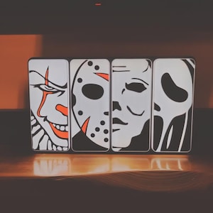 May include: Four white rectangular phone cases with black and orange graphic illustrations of horror movie characters. The characters include a clown, a hockey mask, a blank face, and a ghost face.