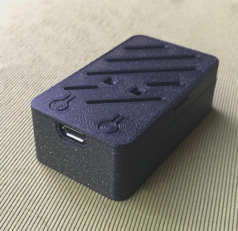 May include: A small, rectangular black device with a textured surface and a USB port. The top features a series of parallel grooves and decorative symbols. The device appears to be 3D-printed.