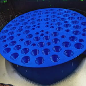 Puede incluir: Un objeto circular azul brillante con numerosos agujeros espaciados uniformemente. El objeto parece estar impreso en 3D y tiene una superficie texturizada. Podría ser un soporte o organizador para objetos pequeños. El objeto está sobre una superficie reflectante.