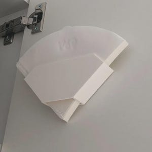 May include: A white, angular, 3D-printed object, possibly a shelf or organizer, mounted on a gray surface. The object has a curved top and a rectangular section. A metal hinge is visible in the upper left corner.