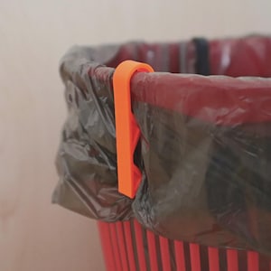 Op de afbeelding: Een oranje plastic vuilniszakclip die een zwarte vuilniszak aan een rood-zwarte gestreepte vuilnisbak bevestigt. De clip heeft een eenvoudig, functioneel ontwerp en zorgt voor een stevige grip op de zak.