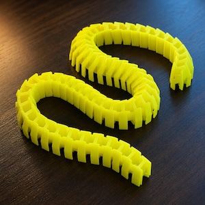 May include: A bright neon yellow articulated cable carrier chain featuring a series of interlocking rectangular segments. The flexible plastic structure is arranged in an S-shaped curve, showcasing a repeating modular design with open side links.
