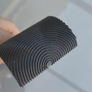 May include: A rolled, semi-circular object with a textured, ridged surface. The material is a dark gray color, with concentric, curved lines creating a pattern. The object appears to be made of a flexible material.