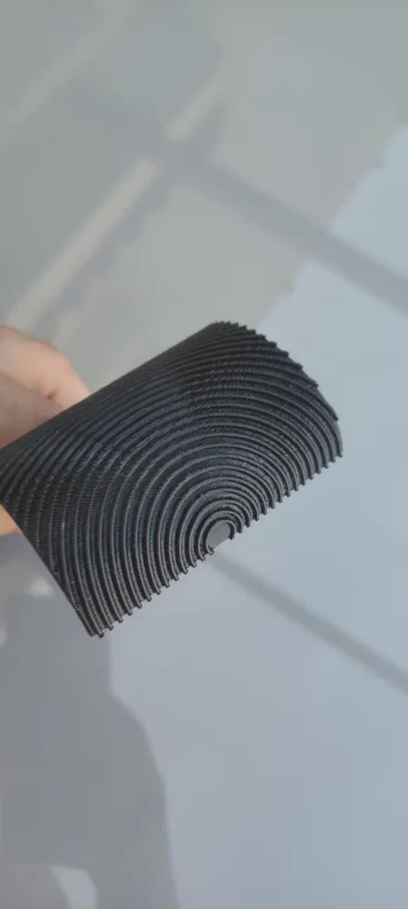 May include: A rolled, semi-circular object with a textured, ridged surface. The material is a dark gray color, with concentric, curved lines creating a pattern. The object appears to be made of a flexible material.