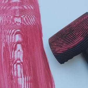 May include: A close-up of a red and black patterned roller tool next to a textured pink paint application. The roller has a black handle and a curved, ridged head. The paint application is on a white surface.