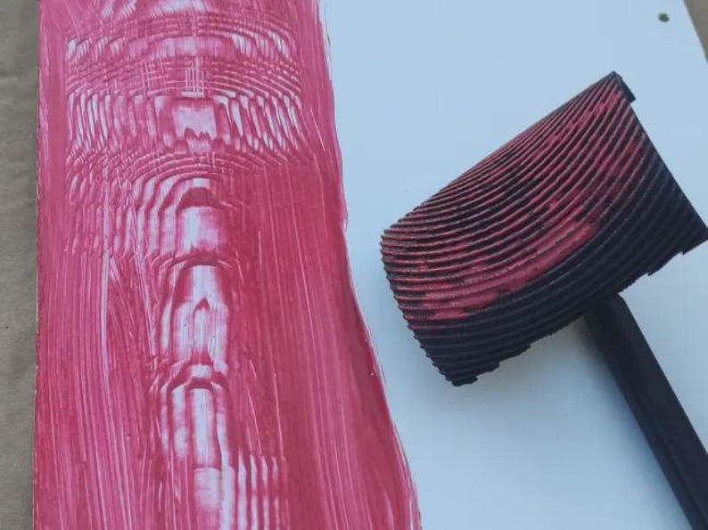 May include: A close-up of a red and black patterned roller tool next to a textured pink paint application. The roller has a black handle and a curved, ridged head. The paint application is on a white surface.