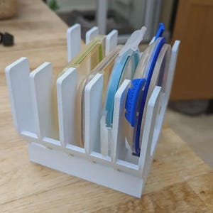 May include: A white plastic storage rack with multiple vertical slots designed to hold various circular container lids. The rack features a sturdy base and upright dividers, currently holding several translucent lids with blue and yellow accents.