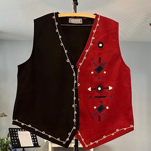 May include: A black and red vest with white trim and geometric embroidery. The vest has a button closure and is hanging on a stand.