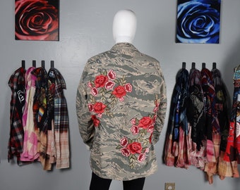May include: A camouflage jacket with red embroidered roses on the back. The jacket is on a mannequin in front of a wall with two paintings of roses and a rack of shirts.