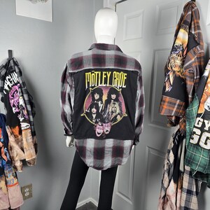 以下が含まれることがあります： Mötley Crüeのバンドが描かれた黒いグラフィックが背中にある、赤、黒、グレーのチェック柄フランネルシャツ。 他のグラフィックシャツが壁に掛かっています。 シャツはマネキンにあります。