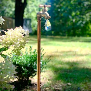 May include: A gold-colored garden faucet with a crystal prism hanging from the spout. The faucet is mounted on a copper pipe and is surrounded by green foliage.