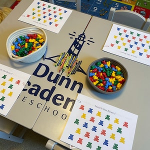 May include: A table with four worksheets that say "Match the Bears!" with colourful bear images. There are two bowls of colourful bear counters, crayons, and a sign that says "Dunn Academy Preschool".