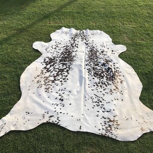 May include: A white cowhide rug with brown spots. The rug is laid out on a green grassy surface.
