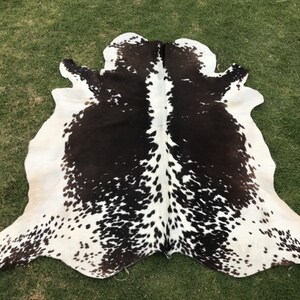 Könnte beinhalten: Ein schwarz-weißer Kuhfellteppich mit einem gefleckten Muster. Der Teppich liegt auf einer grünen Grasfläche.