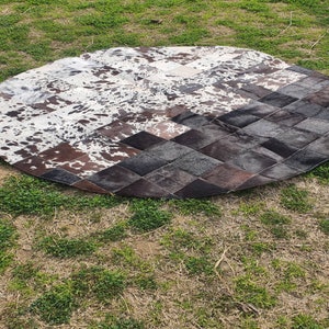 Może przedstawiać: Okrągły dywan z patchworku ze skóry krowiej w kolorze czarnym, brązowym i białym. Dywan wykonany jest z różnych kawałków skóry krowiej zszytych ze sobą, tworząc unikalny wzór.