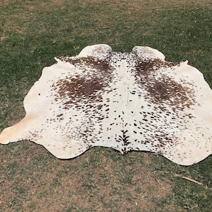 May include: A white cowhide rug with brown spots. The rug is laid out on a green grassy surface.