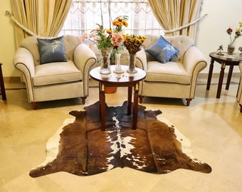 May include: A brown and white cowhide rug with a light brown, cream, and white color scheme. The rug is laid out on a light beige tiled floor in a living room setting with two white armchairs and a wooden coffee table.