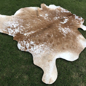 Pode incluir: Um tapete de pele de vaca marrom e branco com uma forma natural irregular. O tapete tem um fundo marrom claro com manchas brancas.