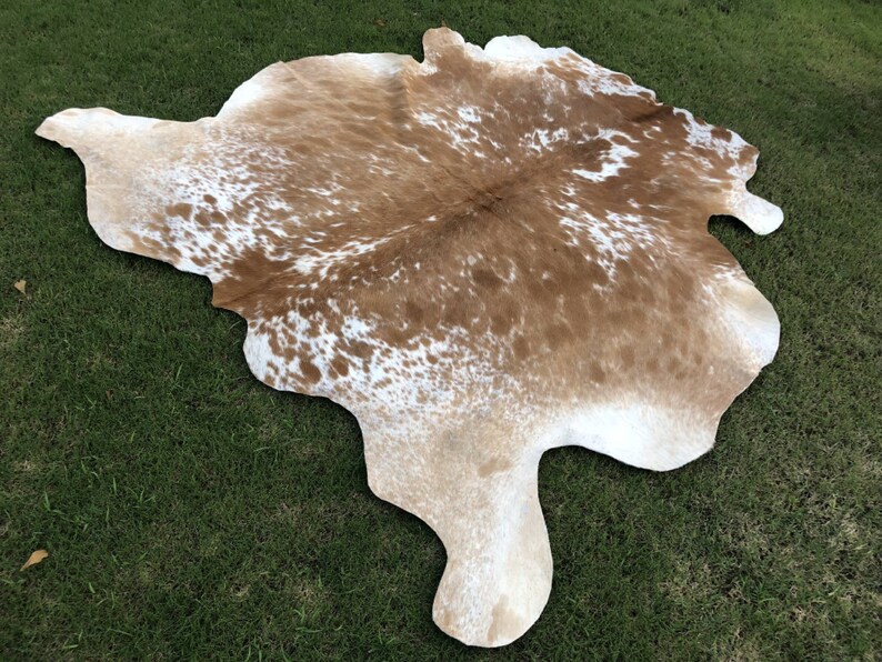 Pode incluir: Um tapete de pele de vaca marrom e branco com uma forma natural irregular. O tapete tem um fundo marrom claro com manchas brancas.