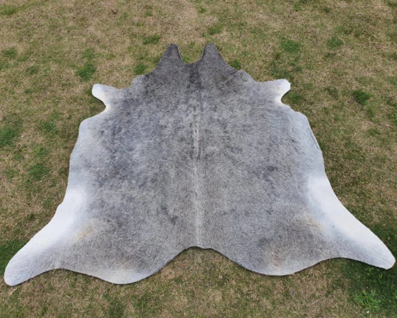 May include: A gray and white cowhide rug with a natural, spotted pattern. The rug is laid out on a green grassy surface.