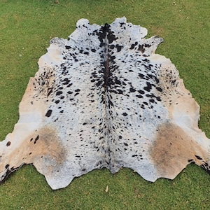 May include: A cowhide rug with a white, black, and brown spotted pattern. The rug is laid out on a green grassy surface.