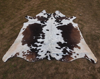 May include: A white and brown cowhide rug with black spots. The rug is laid out on a grassy surface.