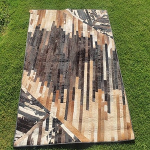 May include: A patchwork rug made of different colored cowhide pieces. The rug is rectangular and has a geometric pattern. The colors are brown, black, white, and tan.