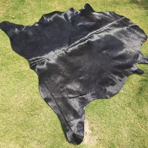 May include: A black cowhide rug with a smooth, shiny texture. The rug is laid out on a green grassy surface.