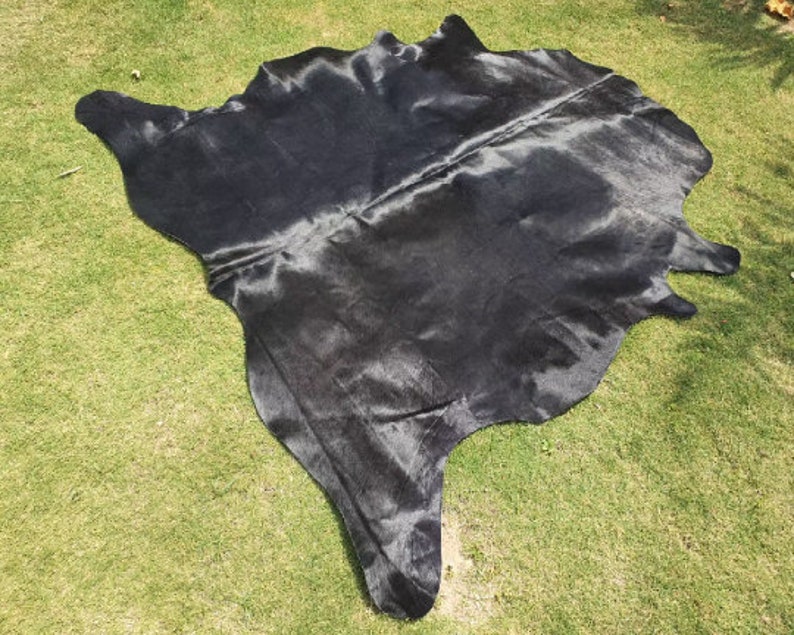 May include: A black cowhide rug with a smooth, shiny texture. The rug is laid out on a green grassy surface.