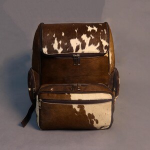 Może przedstawiać: Brązowo-biały plecak ze skóry krowiej z wieloma przegrodami zapinanymi na zamek. Plecak posiada klapę i boczne kieszenie. Torba ma surowy, naturalny wygląd, z mieszanką brązowego i białego futra.