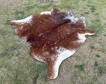 May include: A brown and white cowhide rug with a natural, spotted pattern. The rug is laid out on a green grassy surface.