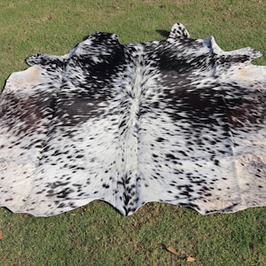 May include: A black and white cowhide rug with a spotted pattern. The rug is laid out on a green grassy surface.