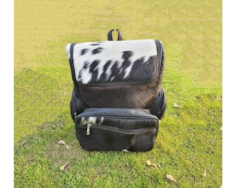 May include: A black and white cowhide backpack with a black leather front pocket. The backpack has a top handle and adjustable straps.