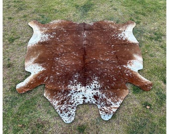May include: A brown and white cowhide rug with a natural, spotted pattern. The rug is laid out on a green grassy surface.