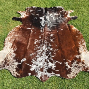 May include: A brown and white cowhide rug with a spotted pattern. The rug is laid out on a green grassy surface.
