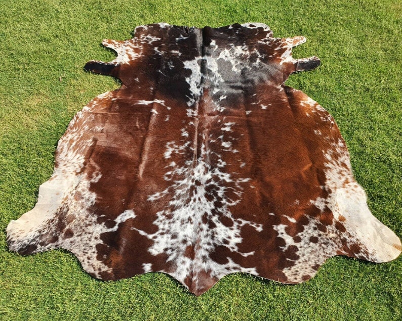 May include: A brown and white cowhide rug with a spotted pattern. The rug is laid out on a green grassy surface.