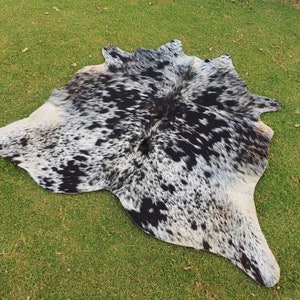 May include: A black and white cowhide rug with a spotted pattern. The rug is laid out on a green grassy surface.