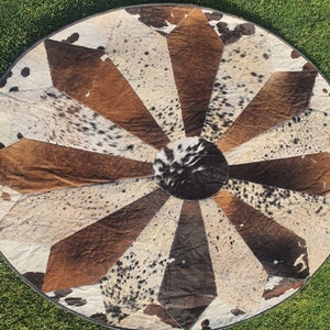 Op de afbeelding: Een rond, veelkleurig koeienhuidkleed met een stervormig patroon. Het kleed is gemaakt van verschillende tinten bruin, zwart en wit.