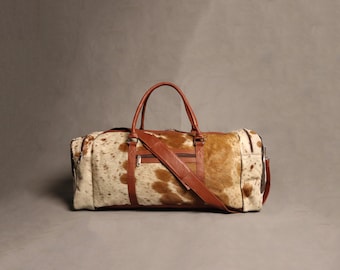 Könnte beinhalten: Eine braune Leder-Reisetasche mit einem weißen und braunen Kuhfellmuster. Die Tasche hat einen oberen Griff und einen Schultergurt.